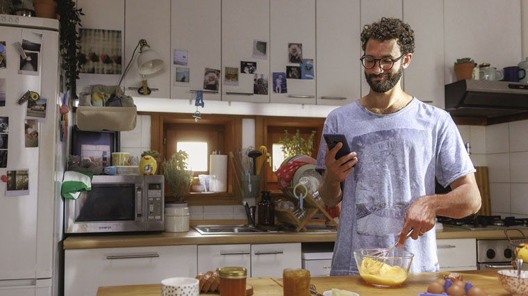 chico en la cocina cocinando con una mano y mirando el móvil con la otra